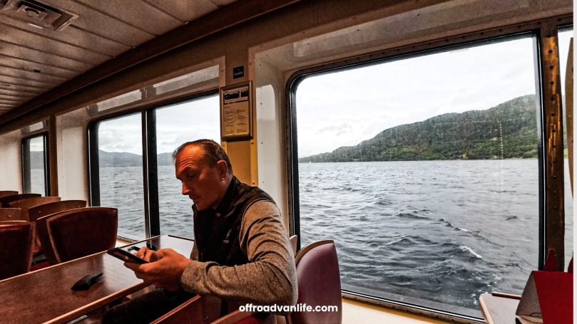 Mann sitzt im Schiff, schaut auf sein Handy, Blick auf den See und die bewaldeten Hügel.