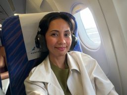 Frau mit langen Haaren und Brille sitzt in einem Flugzeug mit anderen Passagieren im Hintergrund.